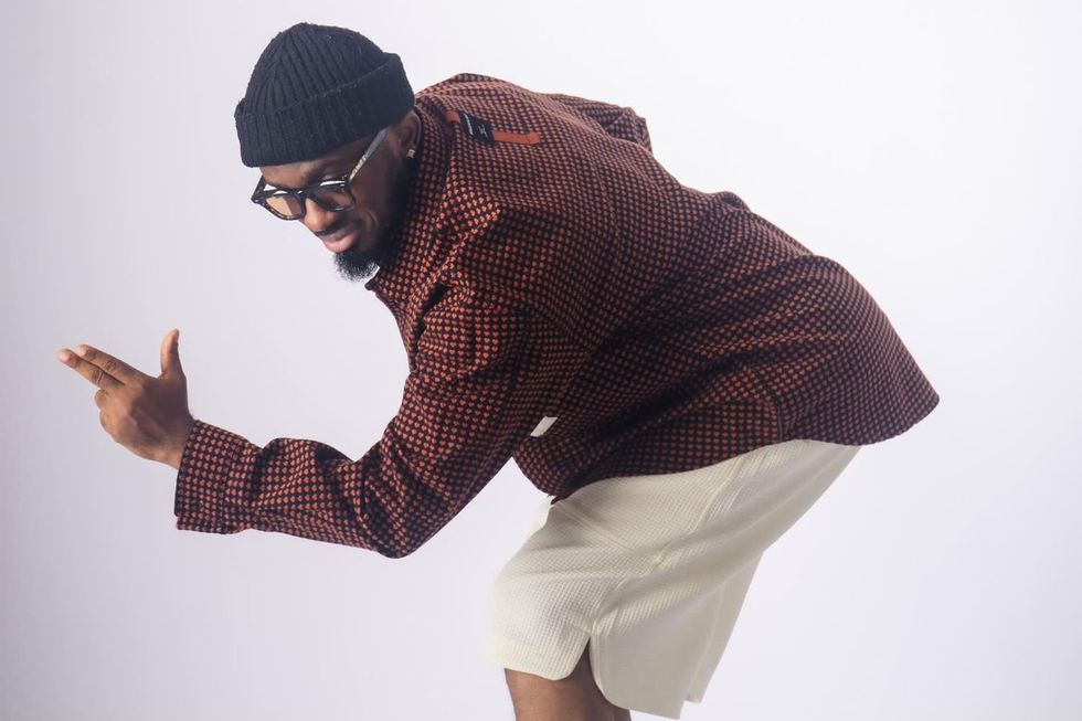 A photo of Fiokee in a knit beanie, patterned jacket, and shorts, striking a playful pose while pointing to the side.