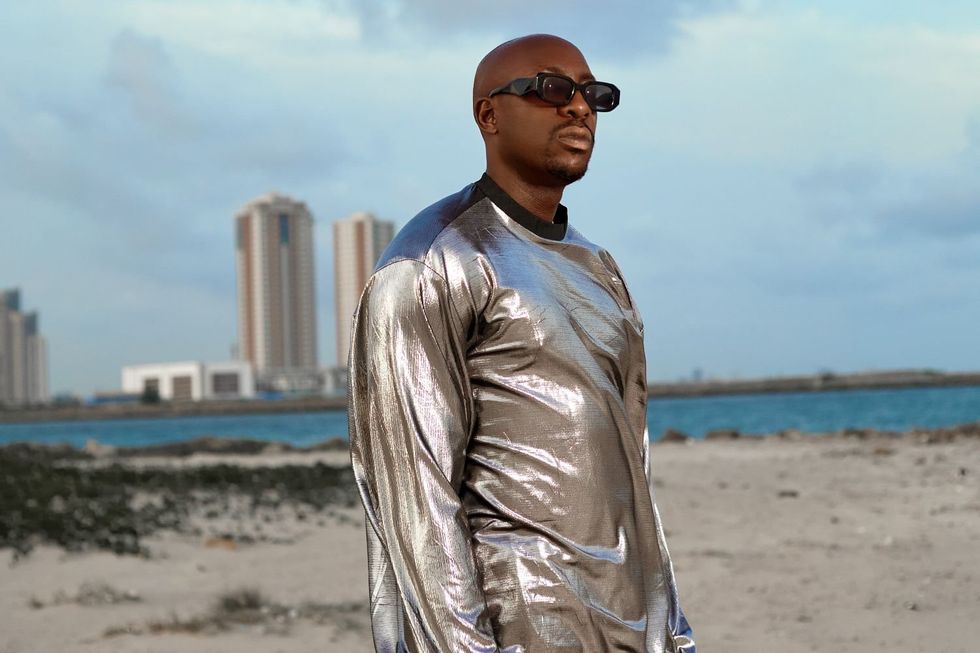 Bien, wearing a shiny silver jacket and sunglasses, stands on a beach.