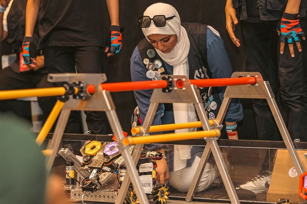 A woman kneels by a robot, appearing to be deep in thought and working with the machine.