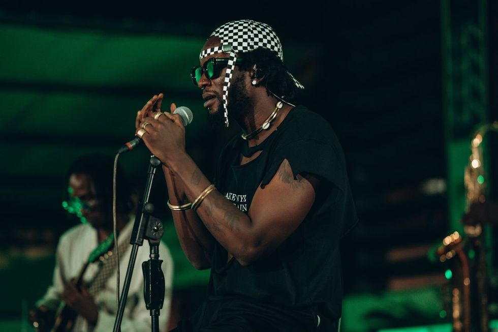 Elijah Kitaka, seated on stage, wearing a black t-shirt, black shorts, and a checkered hat, passionately sings into the microphone.