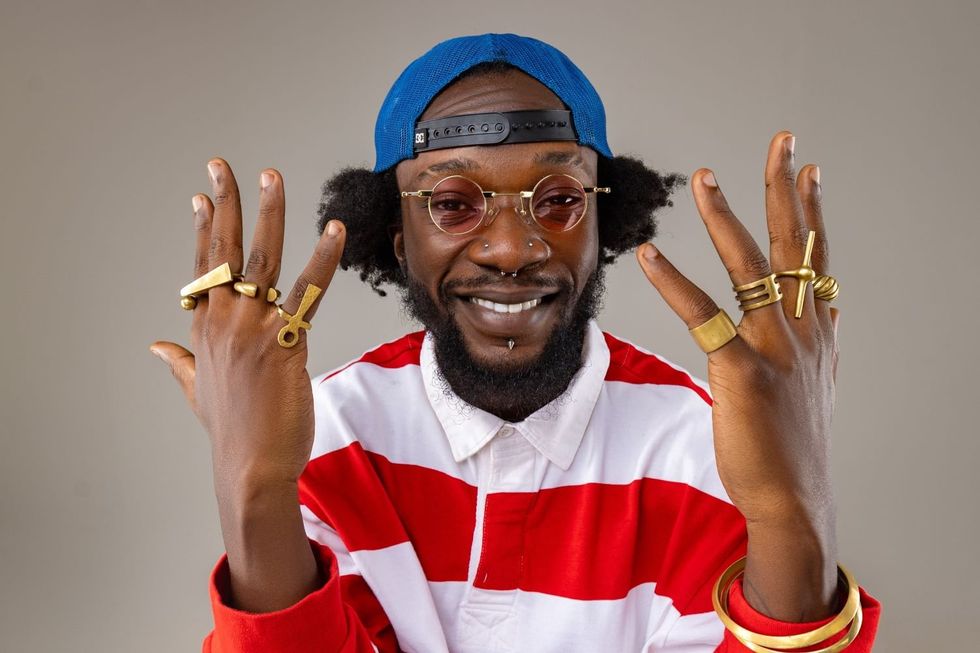 Elijah Kitaka is smiling widely, wearing round glasses, a blue backwards cap, a red-and-white striped shirt and several gold rings and bracelets on both hands.