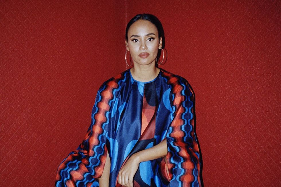 Portrait of Mayra Andrade seated against a red wall, wearing a vibrant blue and red patterned dress.