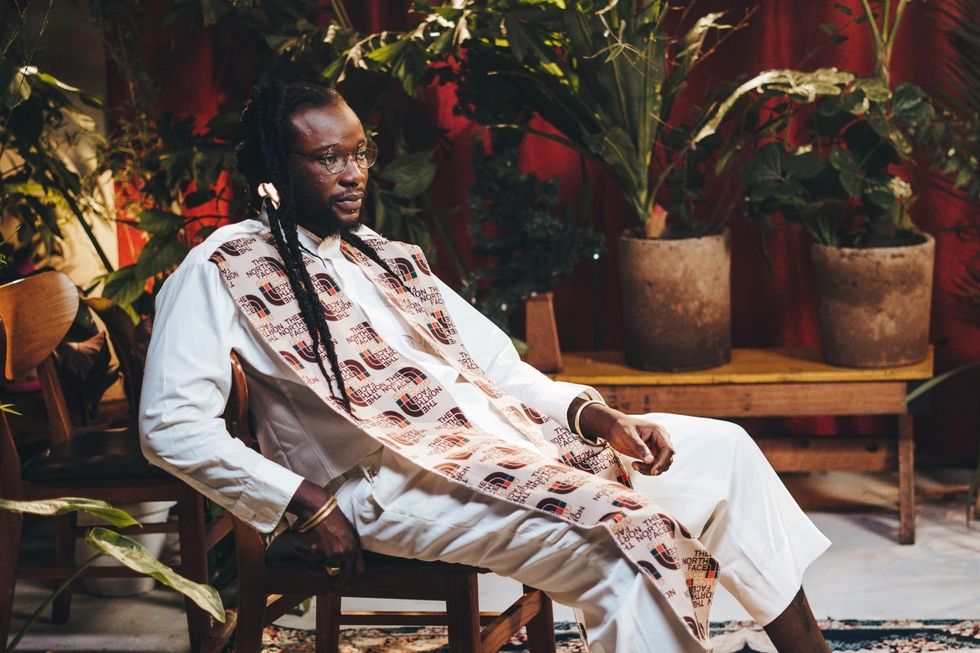 Elijah Kitaka, a Ugandan artist, sits in a chair wearing a white ensemble with a patterned scarf.